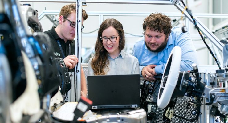 Trzy osoby stoją i rozmawiają przed laptopem. Na pierwszym planie wnętrze samochodu.