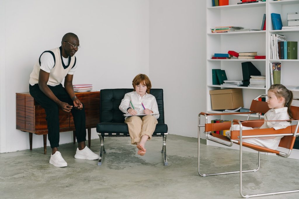 Grafika przedstawiająca mężczyznę rozmawiającego z dziewczynką i chłopcem w pokoju.